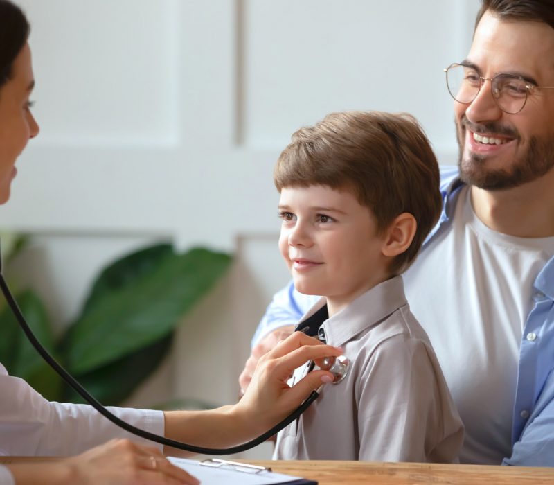 Caring woman doctor hold stethoscope listen to happy small boy patient heart chest in hospital. Young father with little son child do checkup examination at pediatrician in clinic. Healthcare concept.